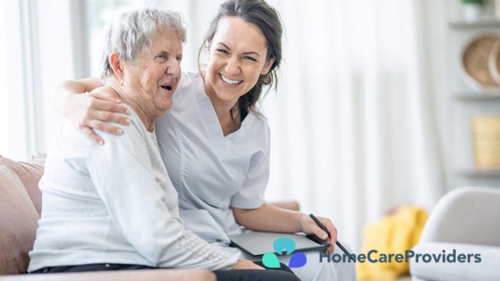 A caregiver smilingly embraces an elderly woman seated on a couch.
